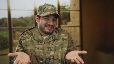 Kamuflaj üniformalı adam avuçlarını kaldırıyor ve boş bina odasında omuz silkiyor; belirsizlik.