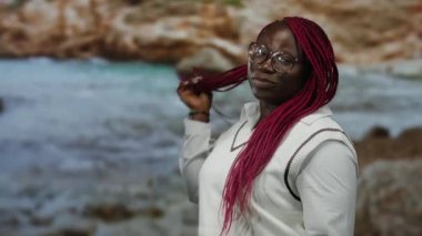 Manzaralı bir sahil kenarında canlı pembe örgüler ve gözlüklerle gülümseyen bir kadın kayalık bir arka plan ve günlük kıyafetlerle neşeli bir kumsal anı yakalıyor..
