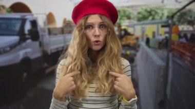 Kabarık saçlı bir kadın hareketli bir caddede kameraya doğru bakıyor, sarışın genç yüz hatları şaşırmış görünüyor, ve gözlüğü ve beresi şehir tarzına uygun..