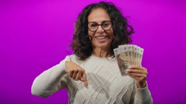 Rusya rublelerini canlı mor bir arka planda tutan, gülümseyen ve aşağıya bakan orta yaşlı bireyler için neşeli bir finansal konsept taşıyan bir kadın..