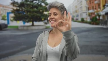 Gri saçlı, günlük kapüşonlu ve kolsuz tişörtlü bir kadın tamam işareti yapıyor ve gülümseyip etrafa bakarken şehir caddesini işaret ediyor..