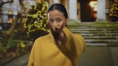 Arka planda festival ışıkları olan bir caddede kendine güveni tam bir kadın jestleri. Sıcak sarı bir süveter giyiyor. Açık hava ortamında dikkatli ve neşeli bir tavır sergiliyor..