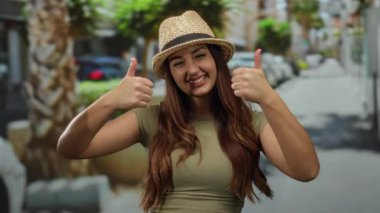 Yeşil tişörtlü ve hasır şapkalı genç kadın, güneşli bir şehir ortamında mutluluk ve pozitifliği ileten palmiye ağaçlarıyla dolu açık bir sokakta başparmağıyla gülümsüyor..