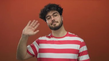 Sakallı, gözlüklü, çizgili tişörtlü, turuncu arka planda sallanan genç adam dostça selamlaşma yapıyor..