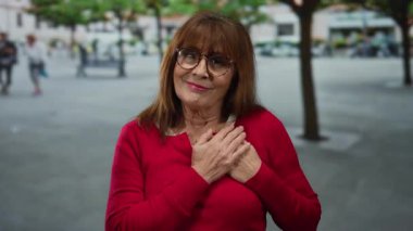 Yaşlı İspanyol kadın göğsünde gözlüklerle gülümsüyor ve şehir sokaklarında kırmızı süveter giyiyor. Ağaçlar bulanık ve arka planda insanlar var..