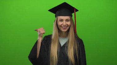 Mezuniyet elbisesi giymiş genç beyaz bir kadın yeşil duvarın arkasına gülümsüyor, mezun olduktan sonra sevinçlerini dile getiriyor..