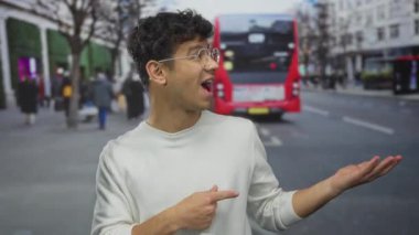 Beyaz kazak giyen genç adam parmak işareti yapıyor ve şehir caddesinde avuçlarını açıyor; heyecan beklentisi.