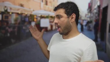 Adam, dükkanlarla ve bulanık yayalarla dolu işlek bir şehir sokağında açık el hareketi gösteriyor; karışıklık.