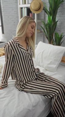 Kadın beyaz yatağa çizgili pijama giyerek uzanıyor ve sarı saçlarıyla başını stüdyoda eğiyor..