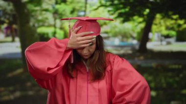 Pembe şapkalı ve cüppeli genç bir kadın dışarıda park ortamında elini güneşli ağaçların altına koyup düşünceli ve düşünceli bir ruh halini yansıtıyor..