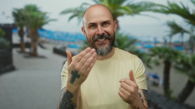 Kel sakallı adam sahilde el kol hareketleri yapıyor. Arka planda palmiye ağaçları ve deniz var. Dostane ve davetkar bir atmosferi var..