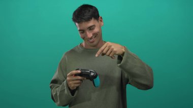 Latin asıllı genç adam elinde Cyan ve Black Game denetçisi gülümseyip yeşil stüdyoyu işaret ediyor; neşe dolu eğlence enerjisi.