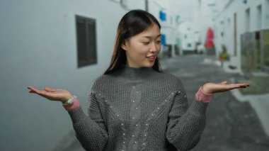 Gri kazaklı Asyalı kadın şehir sokağında ellerini uzatmış düşünceli bir şekilde duruyor..