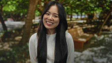 Genç Çinli kadın yemyeşil bir parkta farklı duygular ifade ediyor. Arka planda ağaçlar ve banklar var..