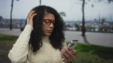 Kıvırcık saçlı ve kırmızı gözlüklü bir kadın, sokakta akıllı telefonu tutarken saçına dokunuyor..