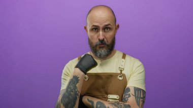 Önlüklü ve siyah eldivenli yakışıklı sakallı adam mor bir arka plana karşı duruyor, düşünceli bir ifade sergiliyor..