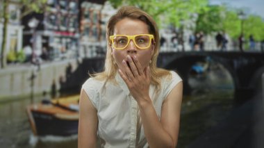 Sarı gözlüklü genç sarışın kadın köprü altındaki kanal teknesinin yanında dudaklarını parmaklarıyla kapatıyor..