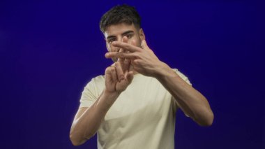 Latin kökenli genç adam mavi stüdyoda elleri ile hashtag jesti oluşturmak için parmaklarını çapraz yapıyor..