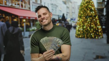 Bize dolar tutan genç adam, arka planda neşeli bir noel ağacı olan şehir sokaklarında gülümsüyor, canlı bir şehir atmosferinde Amerikan paralarını sergiliyor..