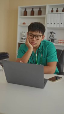 Steteskoplu yeşil önlüklü genç doktor, tıbbi ekipman ve raflarla çevrili modern bir klinikte dizüstü bilgisayar üzerinde çalışırken düşünceli görünüyor..