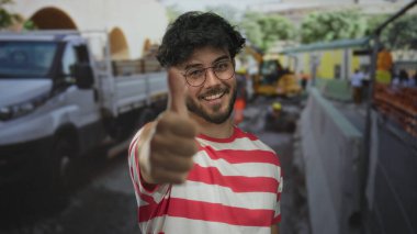 Sakallı ve gözlüklü genç adam inşaat alanında arka planda kamyonla onay veriyor..