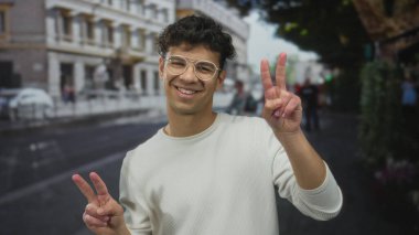 Beyaz süveter giyen ve gözlüklü bir adam iki eliyle sokak köşesinde barış işareti yapıyor. İyimserlik..