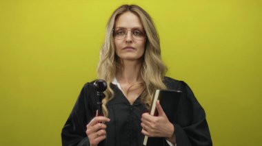 Üniformalı genç sarışın kadın yargıç, canlı sarı arka plan duvarına karşı tokmak tutuyor kapalı alan hukuku kavramına izole edilmiş