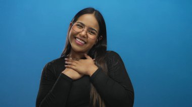 Mavi arka planda kollarını açmış gülümseyen genç bir kadın, gözlük takıyor ve siyah bir bluz giyiyor, izole edilmiş bir stüdyoda neşe ve açıklığın somutlaştırıcısı..