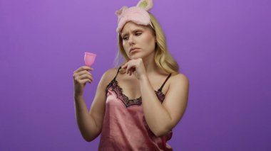 Pembe gecelikli kadın elinde mor arka planda tuhaf bir ifade olan adet bardağı tutuyor.