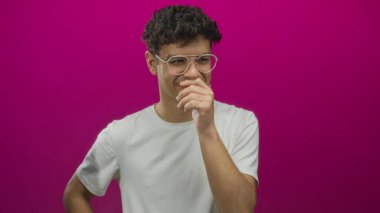 Gözlüklü genç adam kameraya ve ağıza bakan beyaz tişörtlü eflatun stüdyoda gülüyor..