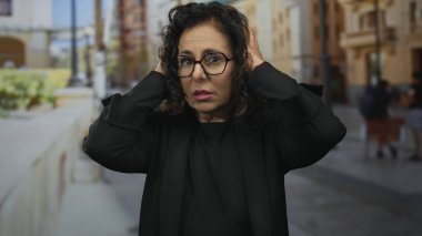 Şehir caddesinde stresi ifade eden, gözlüklü ve siyah elbiseli bir kadın binaların ve bankların bulanık arka planına karşı..