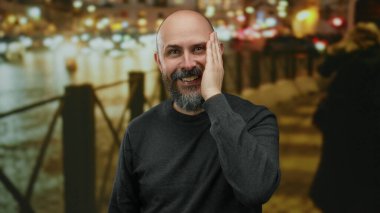 Sakallı bir adam gece şehir sokağında gülüyor. Arka planda bulanık ışıklar var. Canlı ve neşeli bir şehir manzarası yaratıyor..