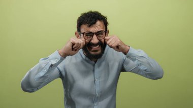 Sakallı ve gözlüklü İspanyol bir adam, sarı arka plandaki üzüntüyü taklit eder. Duygularını yüzüne yakın ifade eder, mavi gömlek giyer..