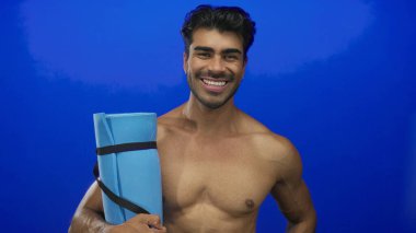 Elinde yoga minderi tutan adam parlak mavi stüdyoda çıplak göğsüne yaslanmış, kendine güveni tam sağlıklı..