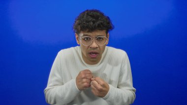 Mavi stüdyoda gözlüklü ve beyaz kazaklı genç adam göğsüne yakın yumruklar kaldırıyor..