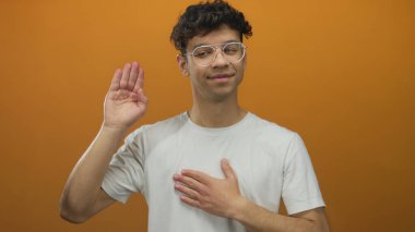 Latin kökenli genç adam sağ elini kaldırıyor ve sol avuç içini turuncu arka plan ile stüdyoya yerleştiriyor; samimiyet yemini onayı.