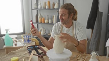 Akıllı telefonu tutan adam seramik vazoyu işaret ederken çömlekçilik stüdyosunda; yaratıcılık odak tutkusu.