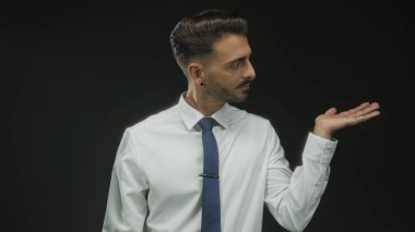 Latin kökenli genç adam, beyaz gömlek ve mavi kravat takıp belirsizliği bir kenara bırakarak siyah stüdyoya avuç içi yayıyor..