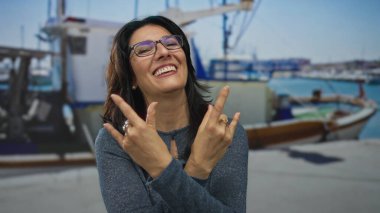 Kadın, deniz kenarındaki teknenin önünde elleri ile taş boynuzları hareketi yapıyor; rock fun metali.