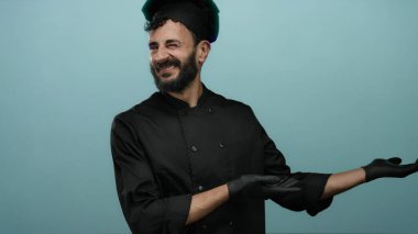 Siyah üniformalı İspanyol aşçı ve eldivenleri mavi arka plana karşı ellerini açmış mutfak bağlamında güvenle gülümsüyor..
