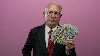 Takım elbiseli kıdemli bir adam elinde dolar banknotları ile pembe bir arka plana karşı, zenginliği ve iş başarısını odaklanmış bir ifadeyle temsil ediyor..