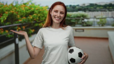 Yeşil arka planı olan bir şehirde elinde futbol topuyla gülümseyen bir kadın açık avuç içiyle dostça bir hareket sergiliyor..