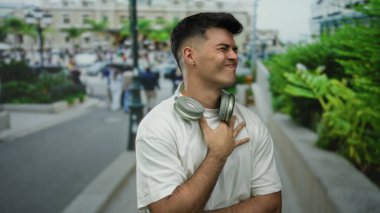 Kulaklıkları boynunda genç bir adam şehir sokaklarında yüz ifadeleri yapıyor. Geçmişi bulanık, beyaz gömlek giyiyor, açık hava manzarası sergiliyor..