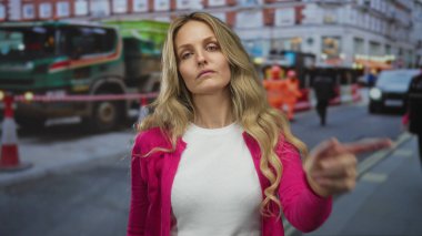 Kendine güveni tam bir kadın inşaat makinelerinin yanında sessiz reddetmeyi işaret ederken sarışın genç bir gözlemci şehir merkezindeki bir cadde boyunca dışarıda duruyor..