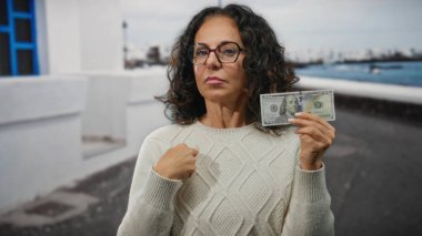 Orta yaşlı bir kadın deniz kıyısındaki bir limanda 100 dolarlık banknota işaret ederek, Amerikan para birimine vurgu yapıyor..