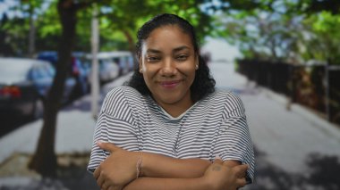 Çapraz kollu bir kadın güneşli bir şehir sokağında park etmiş arabaların ve ağaçların önünde kendine sarılıyor..