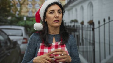 Noel Baba şapkası ve kırmızı kareli önlük giyen İspanyol kadın, park halindeki arabalar ve demir çitlerin yanında, şehir sokaklarında parmak uçlarına dokunuyor..