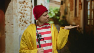 Sarı ceketli ve kırmızı bereli genç adam elinde olta kamışı tutarken tuhaf bir şehir arka planında, düşünceli ve rahat bir havayı çağrıştırıyor..