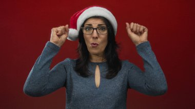 Noel Baba şapkası ve mavi parlak süveter giyen İspanyol kadın canlı kırmızı duvarın önünde kollarını kaldırıyor; bayram neşesi.