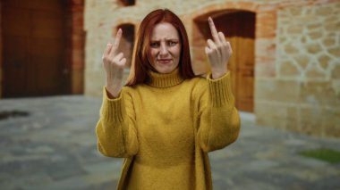 Üniversite kampüsünün dışında el kol hareketi yapan, tarihsel eğitim ortamında meydan okuyan, hardal kazak giyen, kızıl saçlı güzel bir kadın..